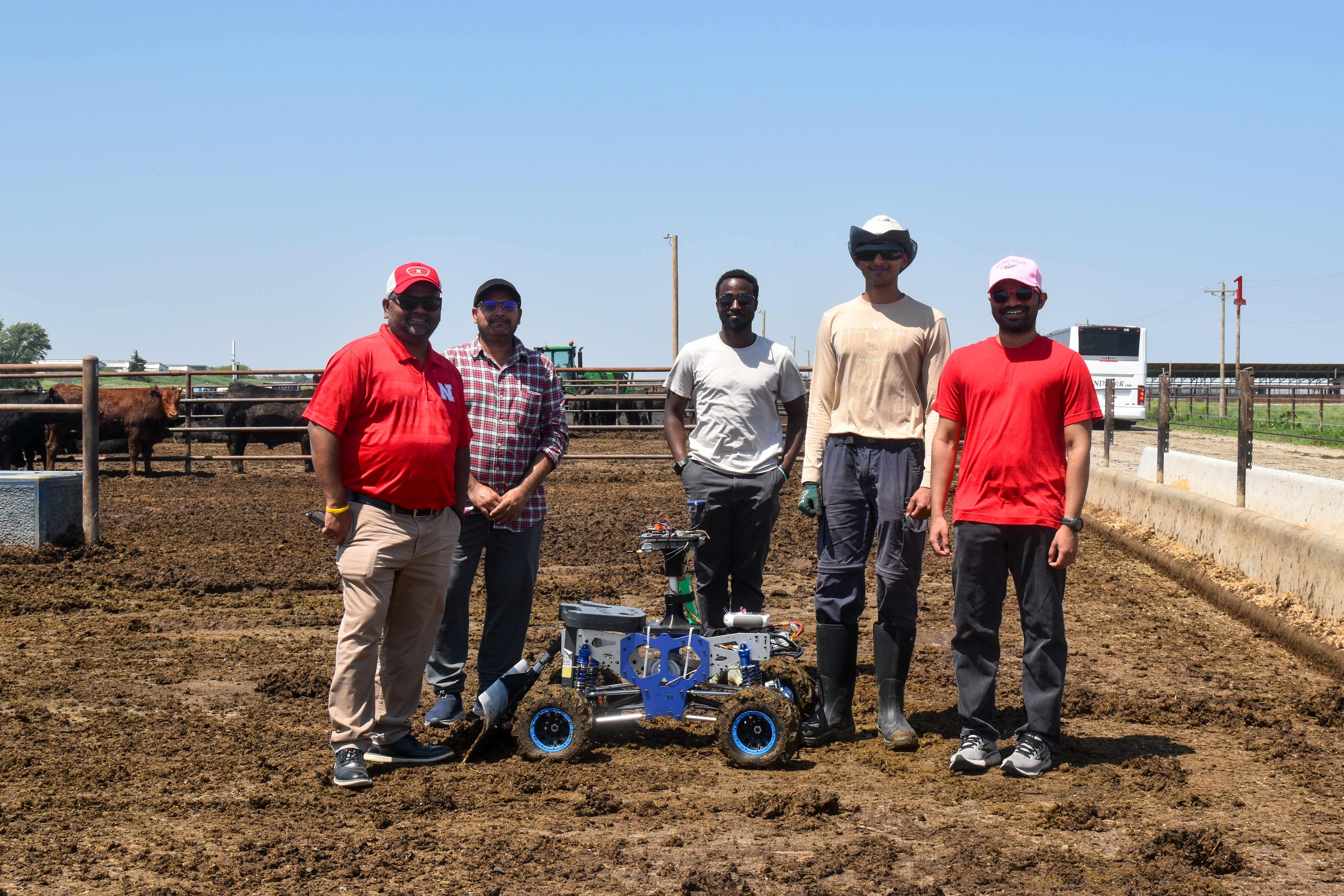 Ag enginnering group demoing prototype for robotic pen scraper in a livestock pen under a clear blue sky.